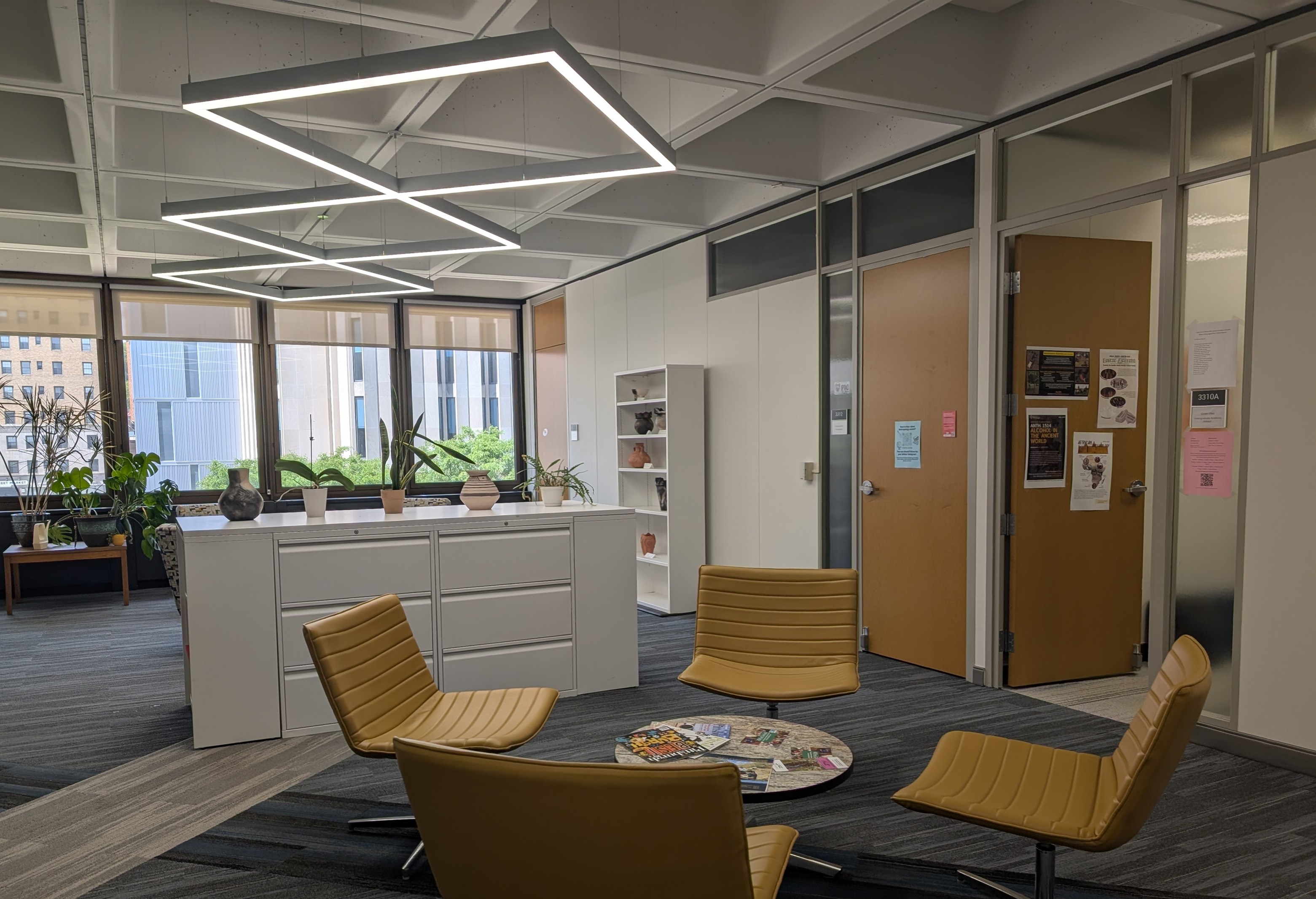 A view of an office space with three doors along the right hand wall and a large sunlight window in the background. Closer to the camera, there are seats with a small table littered with flyers.